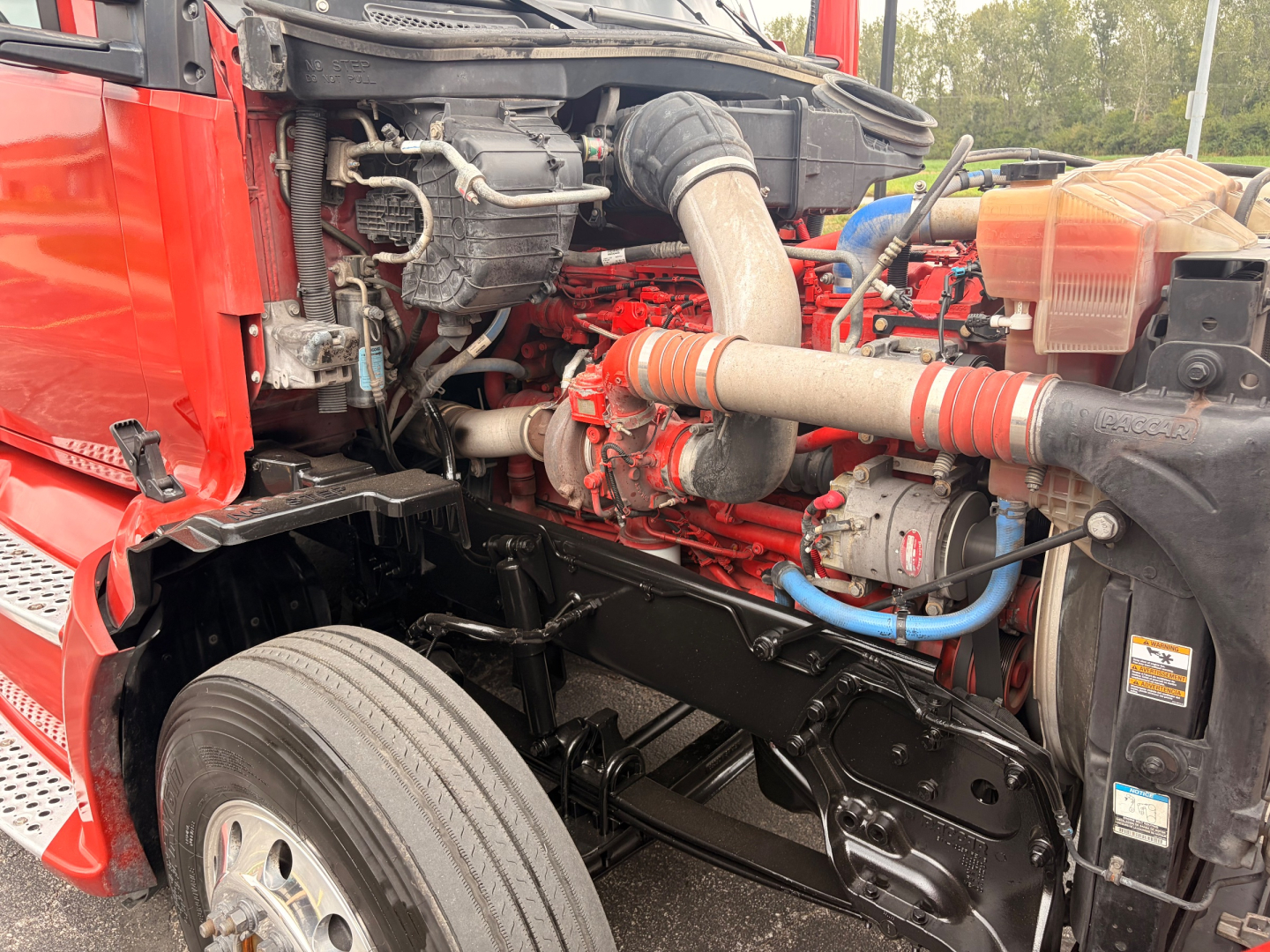Kenworth T680 engine bay showing 12.0L diesel engine with red engine block, large air intake hose, turbocharger, coolant reservoir, and metal frame. Visible components include hoses, wiring, and exhaust piping.