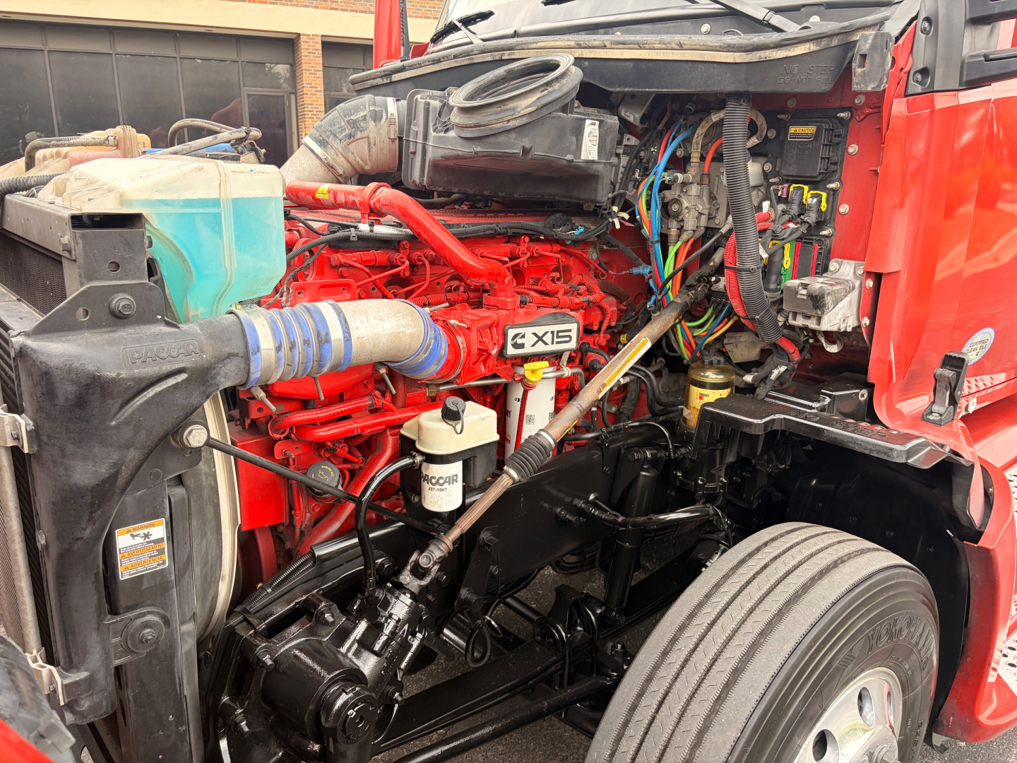 Kenworth T680 engine bay showing X15 Cummins engine with red block, hoses, wiring, and front suspension; visible coolant reservoir, air intake, and various components.