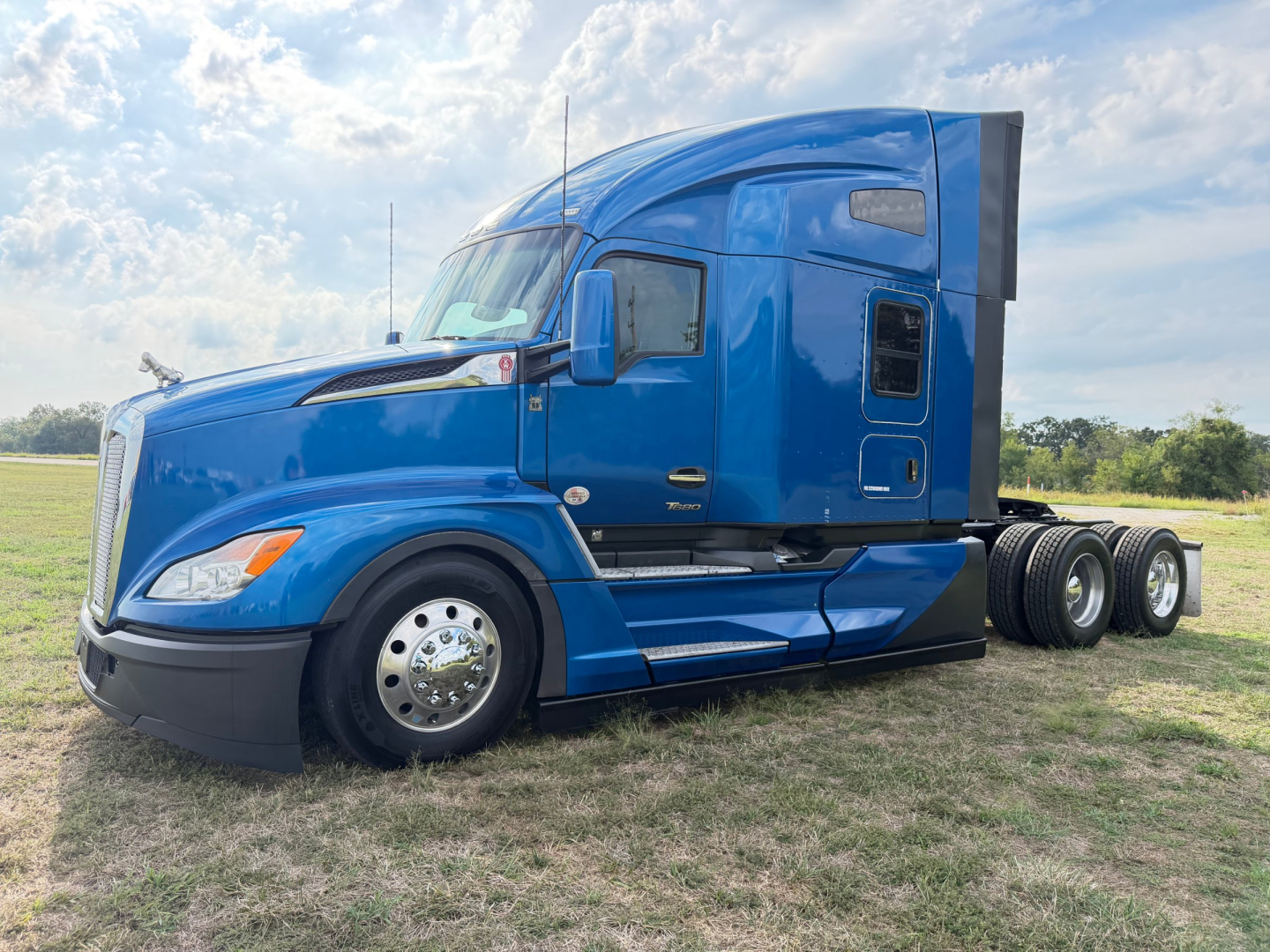 Kenworth T680 tractor in blue with sleeper cab, side view showing front fender, door, and dual rear axle on a grassy lot.