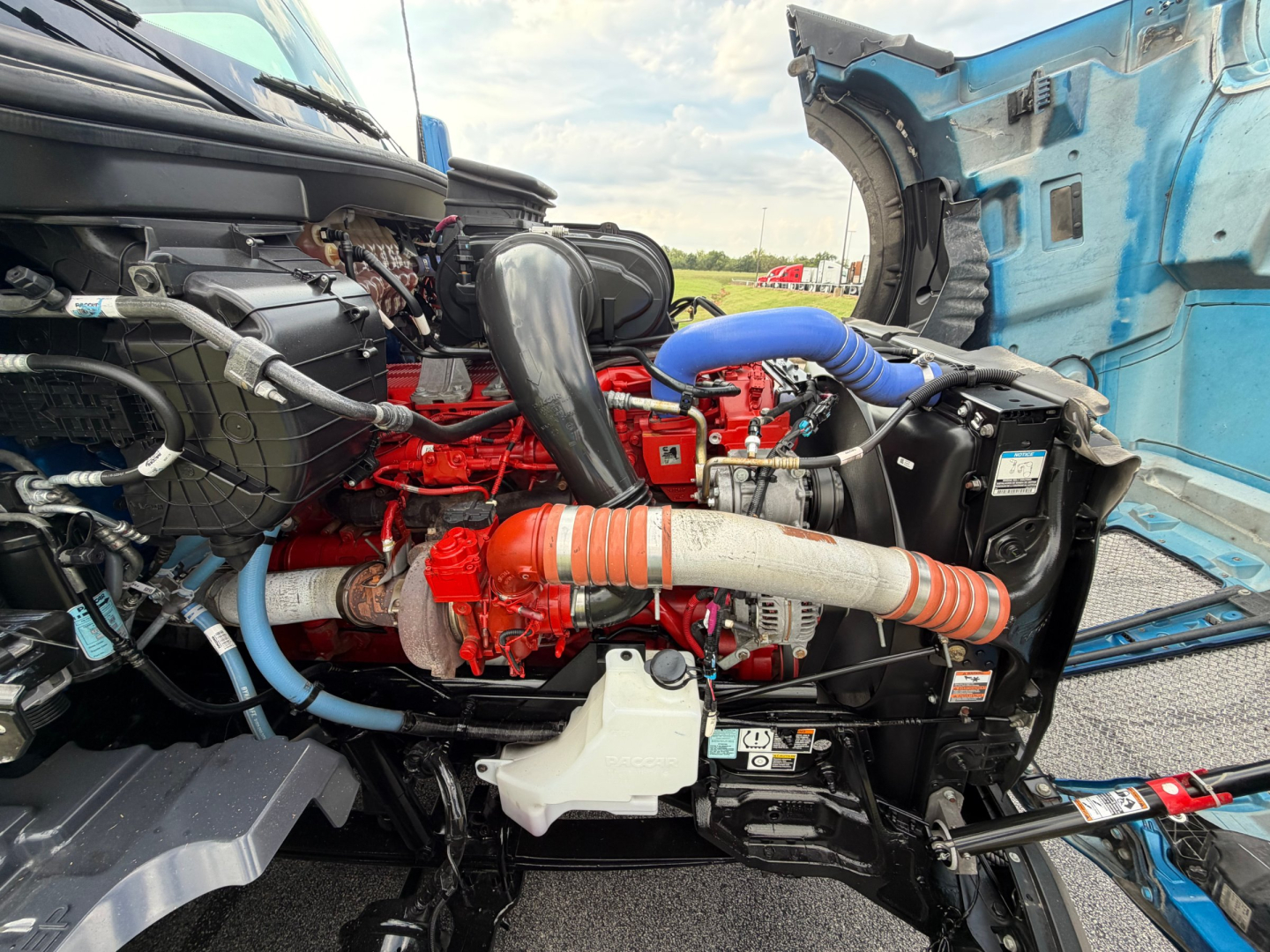 Kenworth T680 engine bay with red engine block, blue coolant hoses, black air intake, and various hoses and components visible.