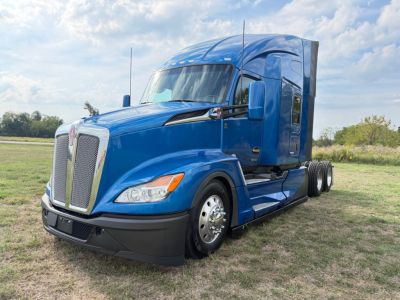 Blue Kenworth T680 truck in a grassy lot, front-left view showing chrome grille, rectangular headlights, side mirrors, and sleeper cab.