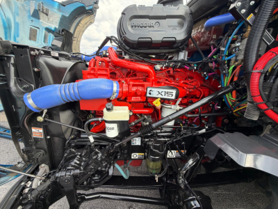 Kenworth T680 engine bay showing bright red Cummins X15 engine with blue coolant hose, air intake, and various hoses and wiring; visible components and labels.