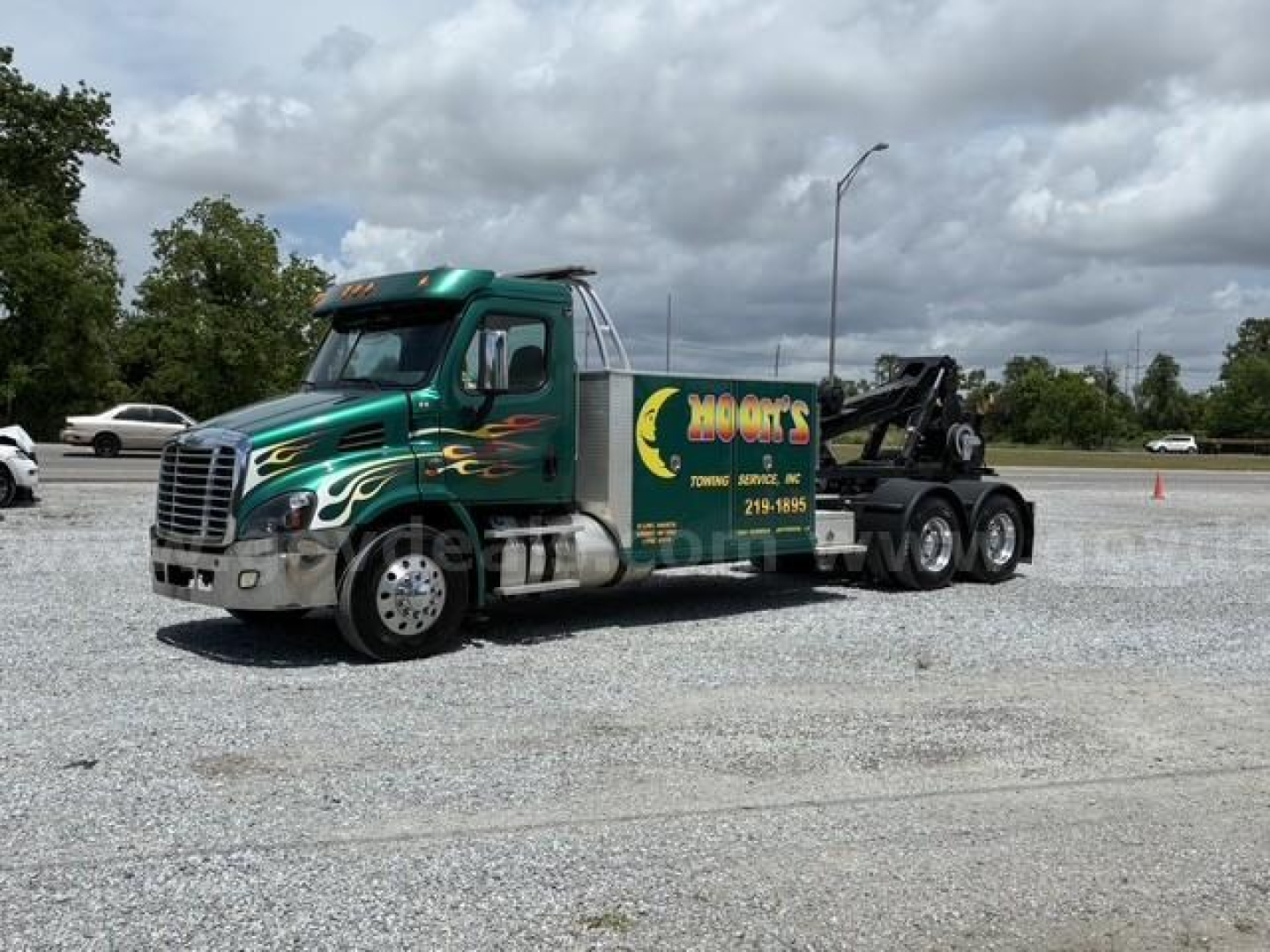 Tow truck with green cab and flame graphics, Freightliner Cascadia 113 tractor-trailer in a parking lot.