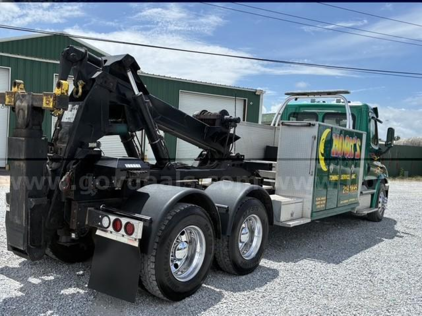 Tow truck with a heavy-duty tow arm and rear axles visible, Freightliner Cascadia 113 in background, green signage on cab side, aluminum toolbox, dual rear wheels.
