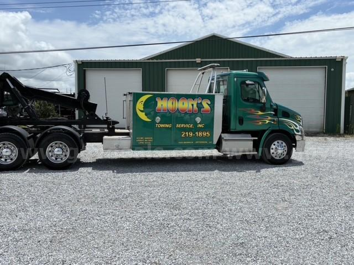 Truck side view showing a green Freightliner Cascadia 113 tow truck with HOON'S Towing Services branding on the cab door and a large steel sleeper behind, parked in front of a green metal building.