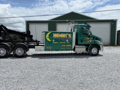 Truck side view showing a green Freightliner Cascadia 113 tow truck with HOON'S Towing Services branding on the cab door and a large steel sleeper behind, parked in front of a green metal building.
