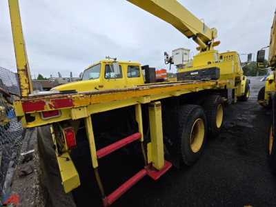 Heavy-duty crane truck in yellow with flatbed and outriggers visible; National Crane boom in high position; rear wheels and chassis show multiple axles; some equipment and another yellow vehicle in background.