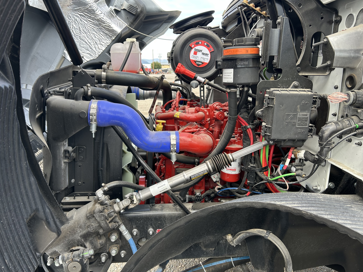 Engine bay of a 2025 Mack MD7 showing red engine block with blue radiator hose, black hoses, wiring, and a circular label on a component near the top.