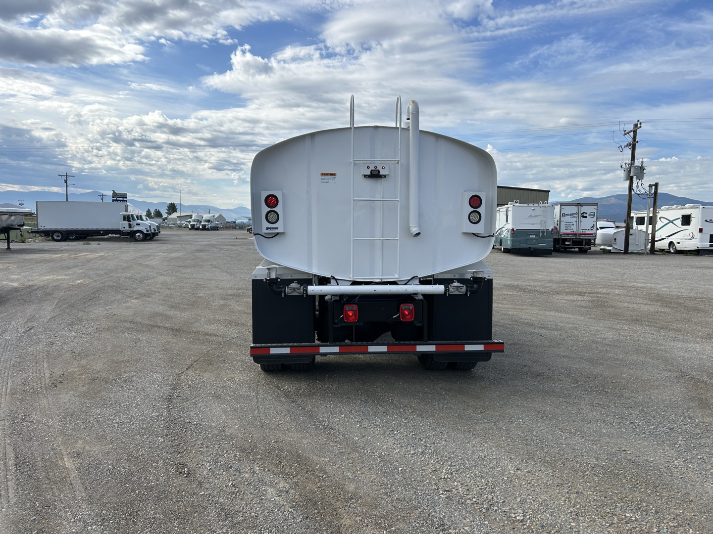 Rear view of a white Mack MD7 tanker truck parked in a lot with gravel ground; tail lights, rear bumper, and ladder visible; clear blue sky with clouds.
