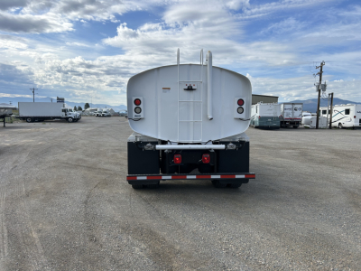 Rear view of a white Mack MD7 tanker truck parked in a lot with gravel ground; tail lights, rear bumper, and ladder visible; clear blue sky with clouds.
