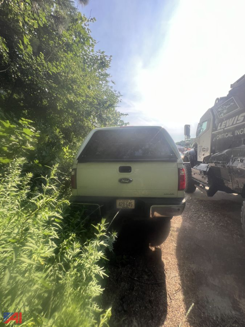 Rear view of a white Ford F-250 Super Duty pickup truck with cap, parked on a gravel road next to a tow truck; vegetation on left.