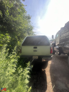 Rear view of a white Ford F-250 Super Duty pickup truck with cap, parked on a gravel road next to a tow truck; vegetation on left.
