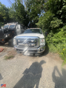 Front view of a white 2015 Ford F-250 Super Duty extended cab pickup truck in a lot, with a chrome grille, bumper, and Ford emblem visible; another tow truck and trees in the background.