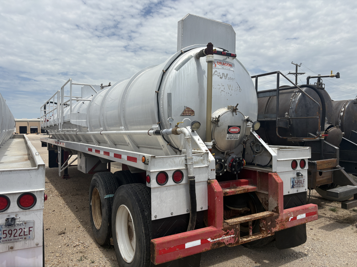 Tank trailer with cylindrical stainless steel tank on dual rear axles, elevated ladder on left, rear bumper with red and white reflective tape, circular access hatch, and a small black diesel tank behind. Visible text: some illegible markin