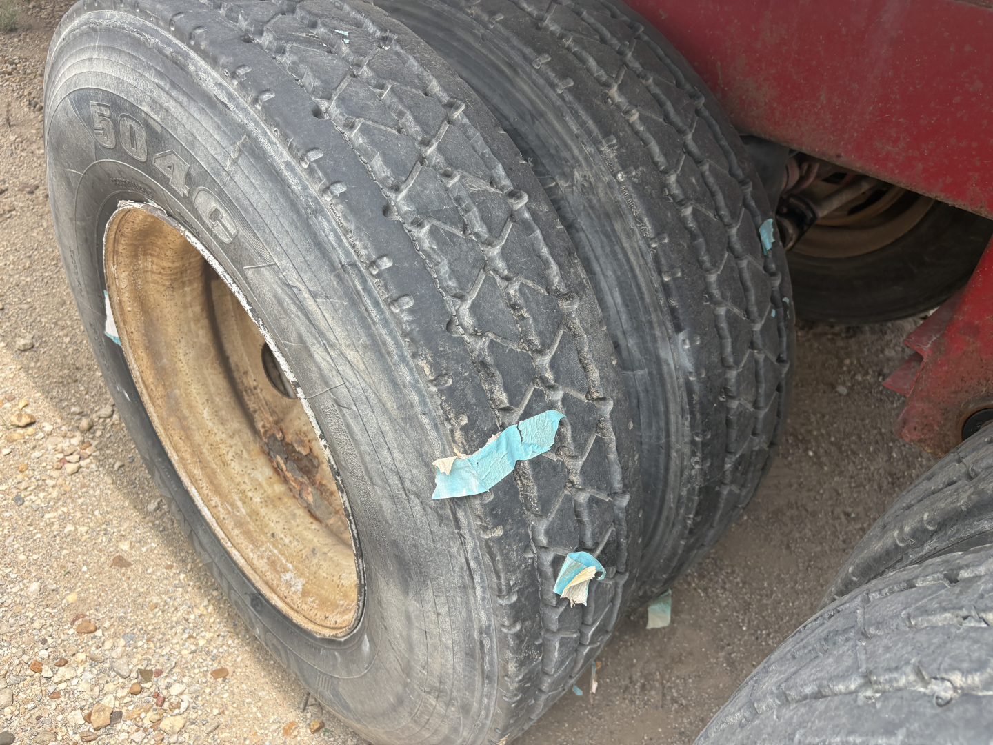 Back wheel of a red trailer showing a weathered tire with visible tread, sidewall markings, and a leather-colored rim. Two pieces of blue tape on the tire. Dusty ground.