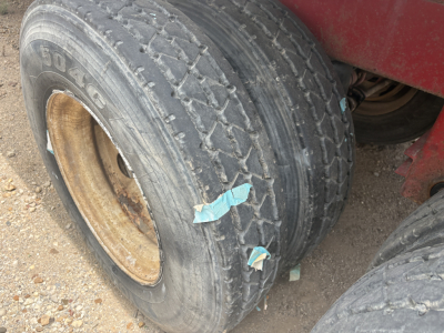 Back wheel of a red trailer showing a weathered tire with visible tread, sidewall markings, and a leather-colored rim. Two pieces of blue tape on the tire. Dusty ground.