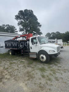 Truck with a red crane grapple on a white Freightliner cab, flatbed body, gravel lot, dual rear wheels, utility equipment in background.