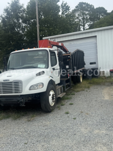 Forklift-mounted grapple on a Freightliner truck in a gravel lot with a white cab and red boom.