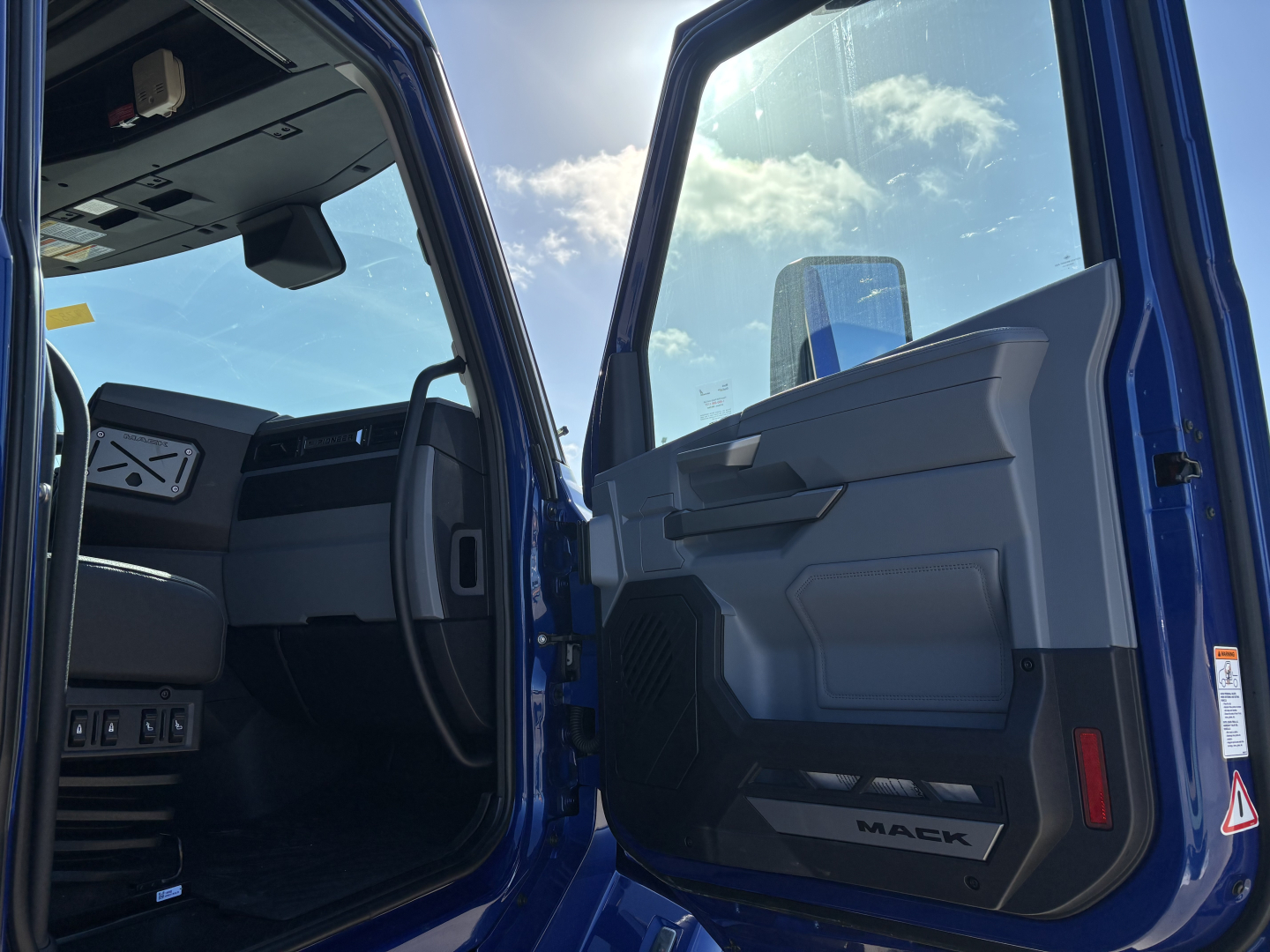 Truck cab interior view of a blue 2027 Mack PR64T76H conventional sleeper, showing open driver door with Mack logo on sill and dashboard area.