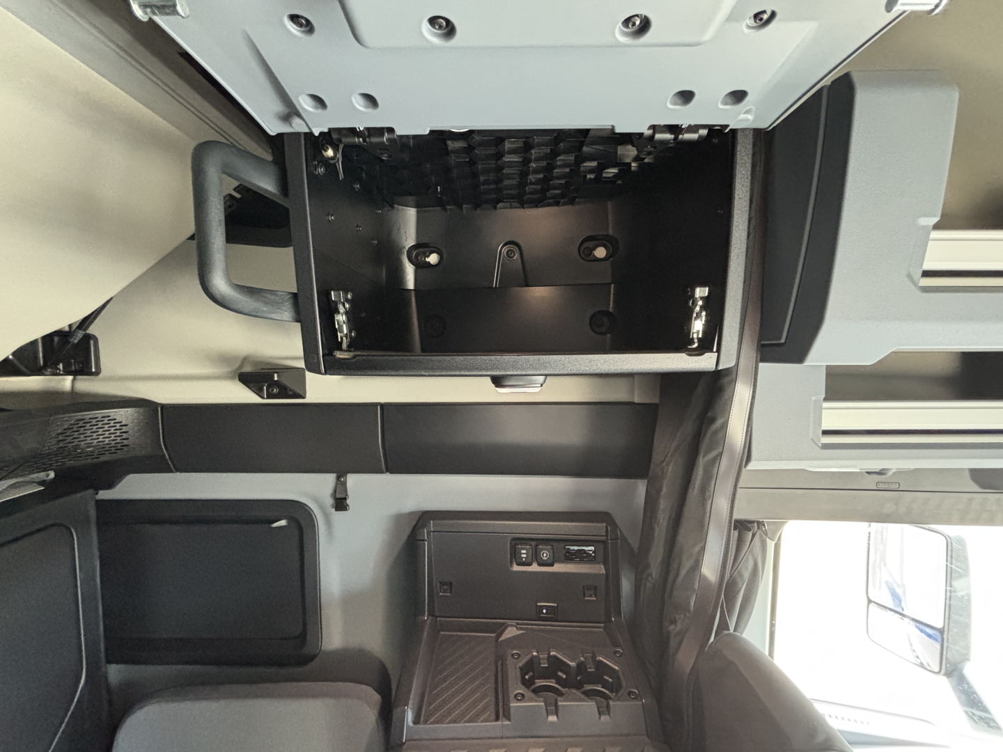 Truck cab interior showing dashboard area and overhead module with empty storage box; center console with switches and cup holders; windows and seating visible.
