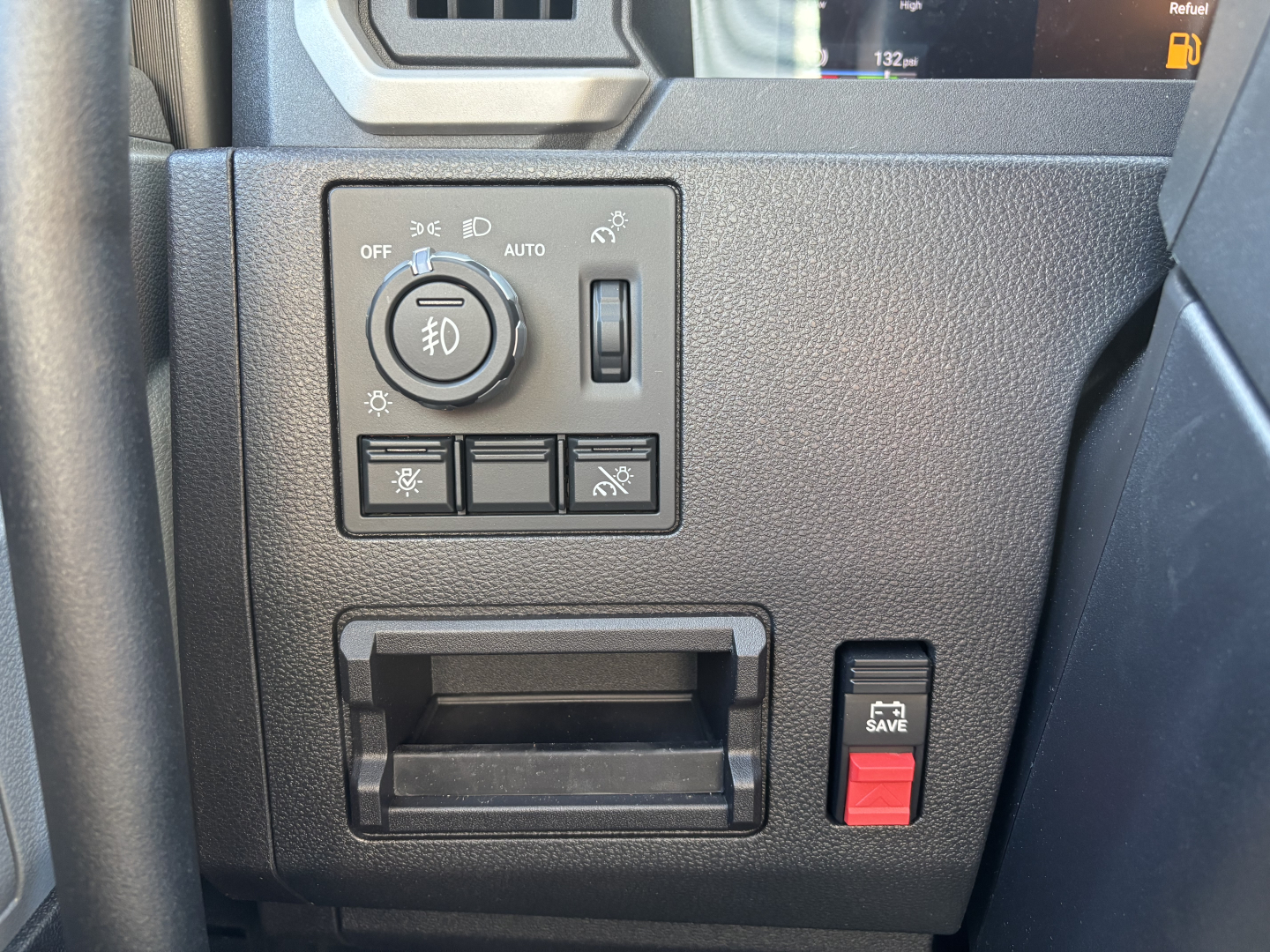 Truck control panel area with headlight dial, fog light switch, and auxiliary buttons; small storage pocket and red hazard-save switch visible.