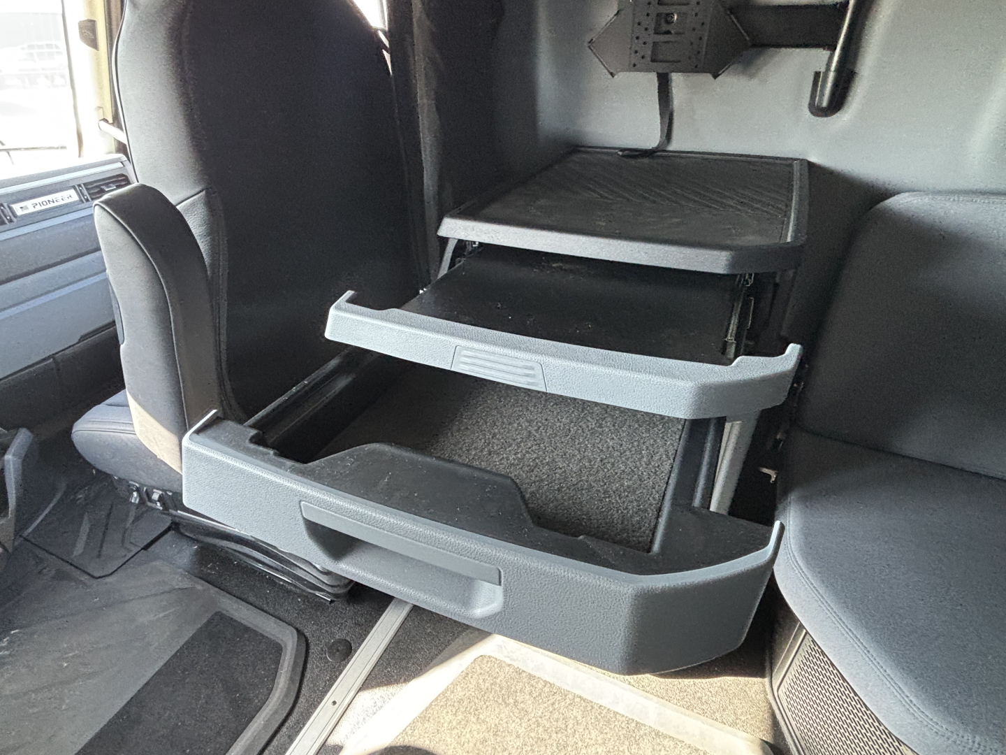 Truck cab interior showing a three-drawer under-seat storage system in the driver area of a Mack PR64T76H.