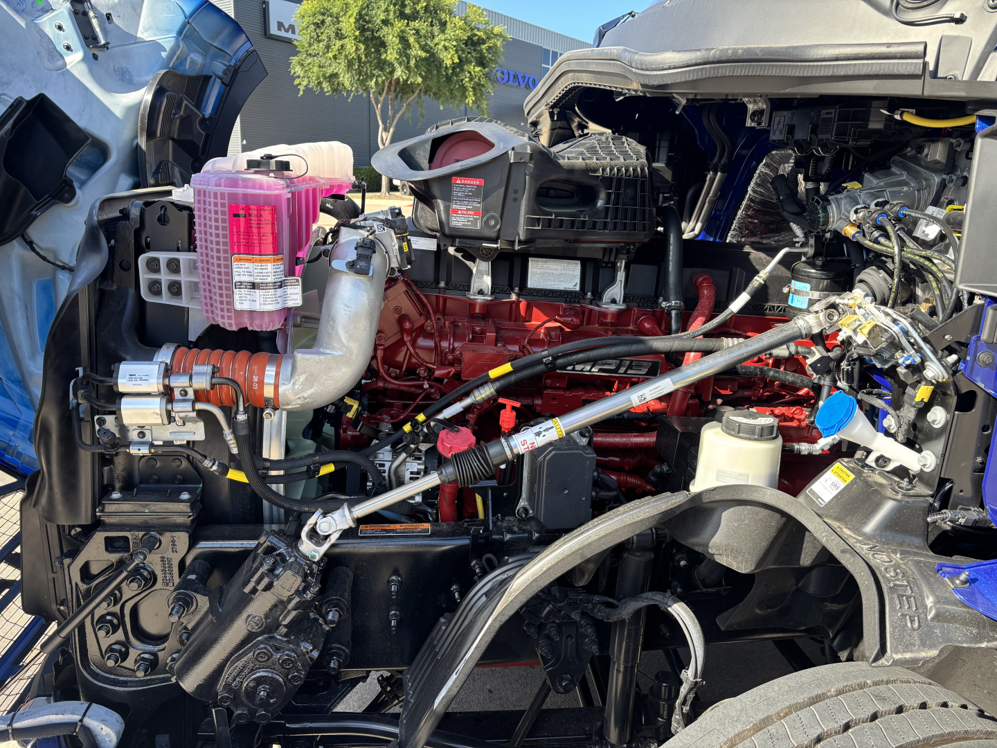 Mack PR64T76H engine bay showing pink coolant reservoir, red engine block, various hoses and the air intake, with fuel and hydraulic lines visible.