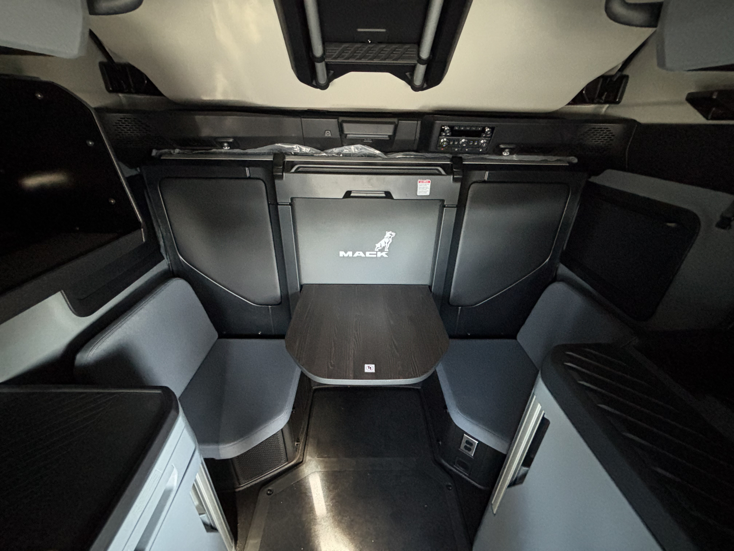 Cab interior of a new 2027 Mack PR64T76H truck showing sleeper area with table, cabinets, and dashboard visible at the front.