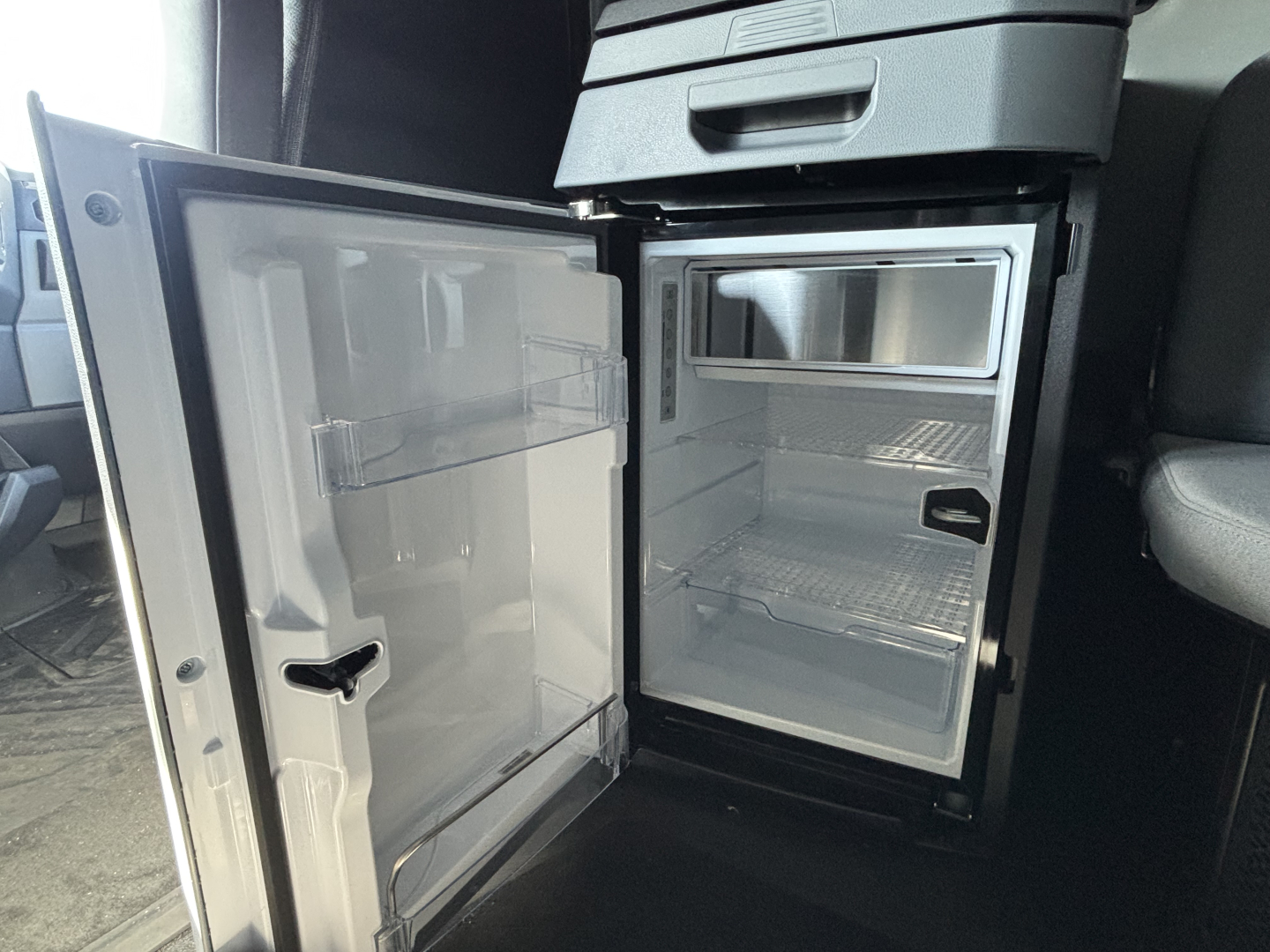 Truck cab interior showing open compact refrigerator/freezer unit inside a Mack PR64T76H sleeper with empty shelves.