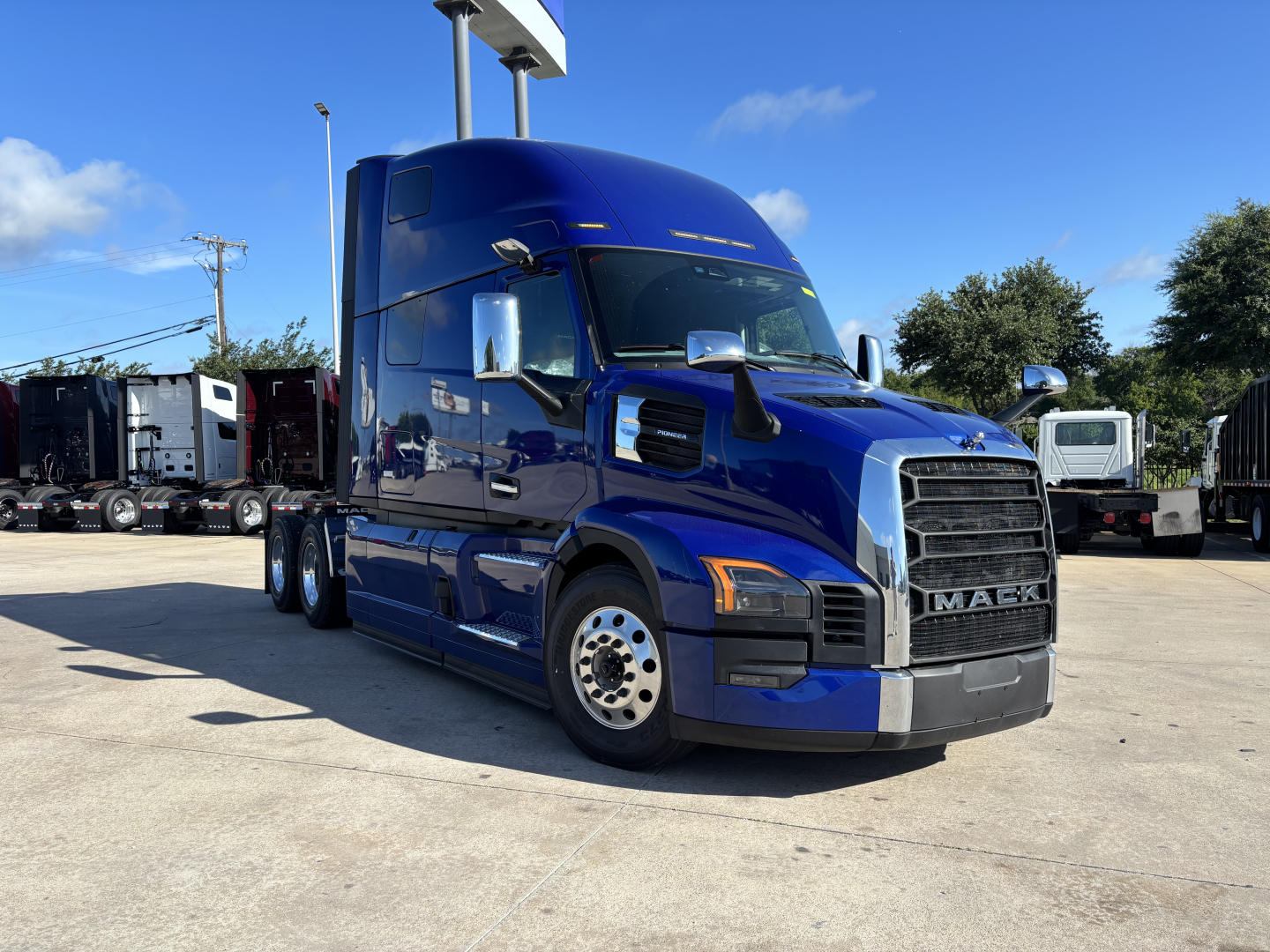 Mack PR64T76H sleeper tractor in blue, front-left view, with grille, headlights, large mirrors, and chrome wheels; dealership lot in background.