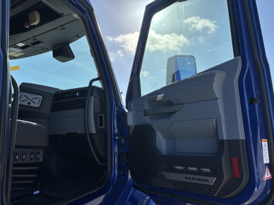 Truck cab interior view of a blue 2027 Mack PR64T76H conventional sleeper, showing open driver door with Mack logo on sill and dashboard area.