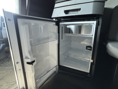 Truck cab interior showing open compact refrigerator/freezer unit inside a Mack PR64T76H sleeper with empty shelves.