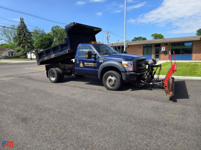 Ford F-450 Super Duty dump truck with plow attachment in a parking lot, dark blue cab, black dump bed, lettering on door, front plow blade in red, clear sky