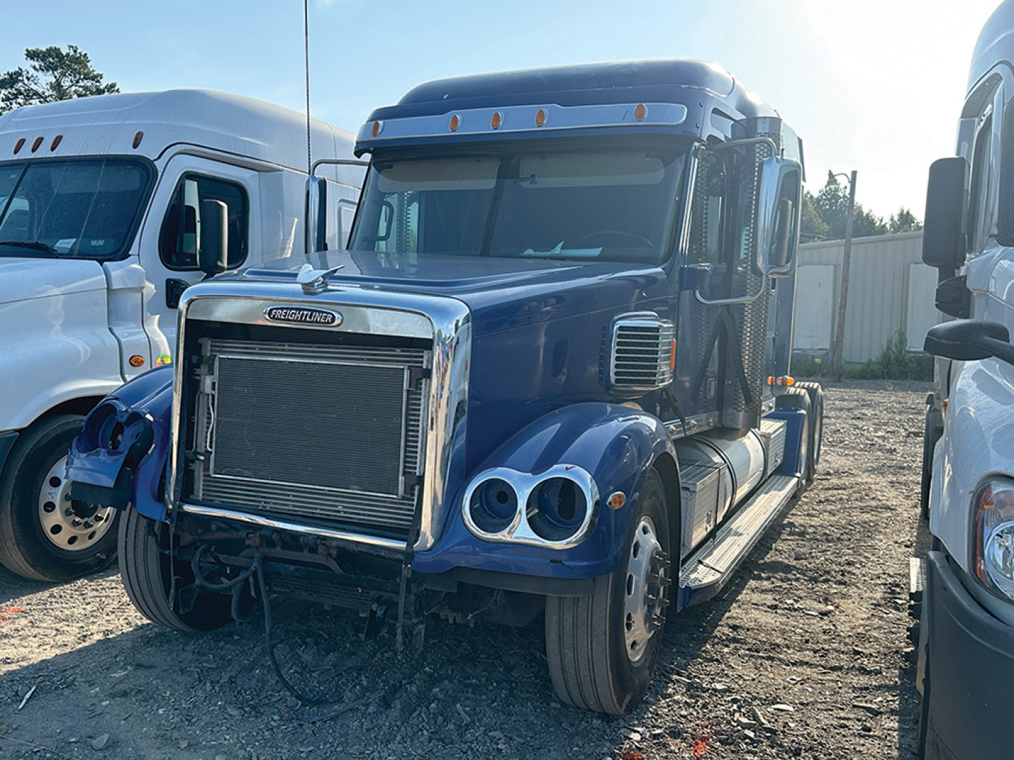 Conventional daycab truck front view showing large chrome radiator grille, dual round headlights, blue cab, and exposed front bumper; Freightliner Coronado styling.