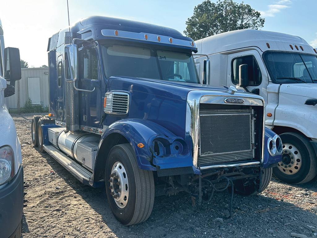 Blue Freightliner Coronado truck cab in foreground with chrome grille and radiator visible; hood area shows absence of bumper; other white truck in background.
