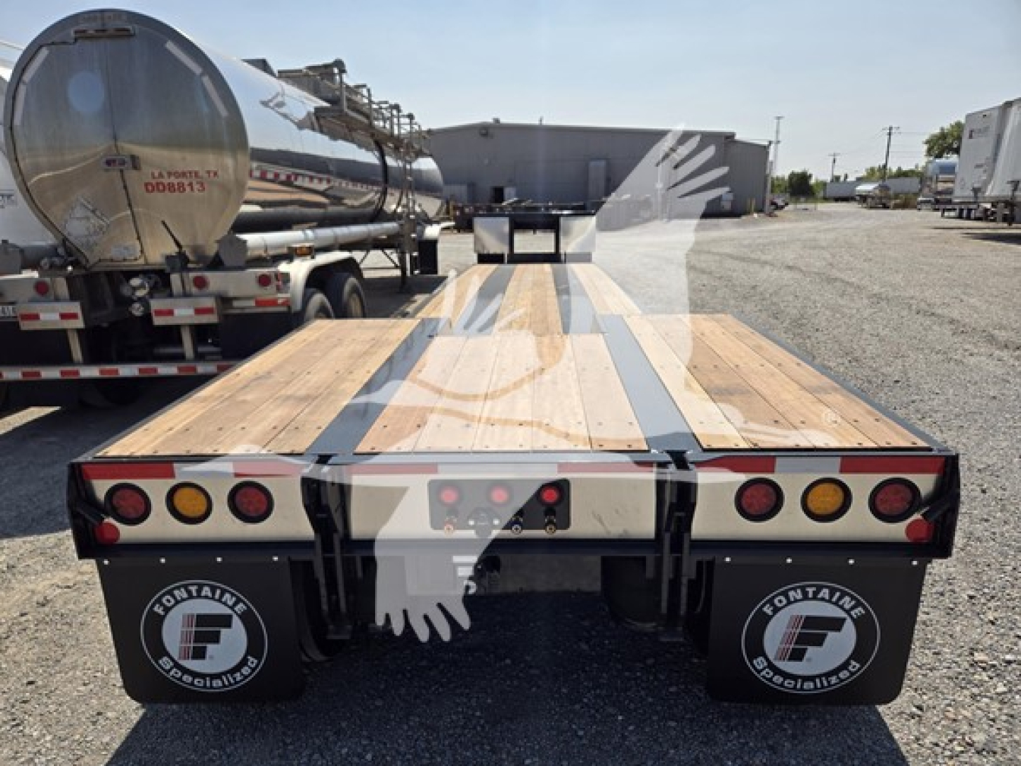 Flatbed trailer deck view from rear showing ramps and taillights, Fontaine Specialists logo mud flaps, wooden deck with center steel support, tanker trailer and yard in background.