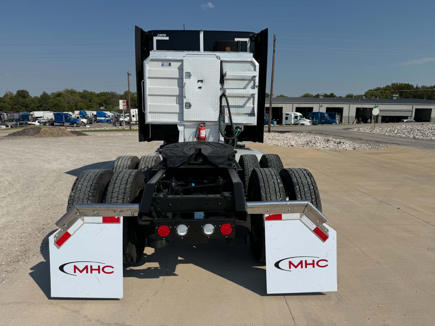 Truck rear view showing fifth wheel, dual rear axles, mud flaps with MHC logo, and metal storage box on a Kenworth T680 chassis.