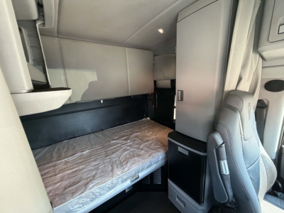 Interior sleeper cab of a 2023 Kenworth T680 showing a single bunk bed with mattress, overhead cabinets, and a small refrigerator/cabinet unit near the passenger seat.