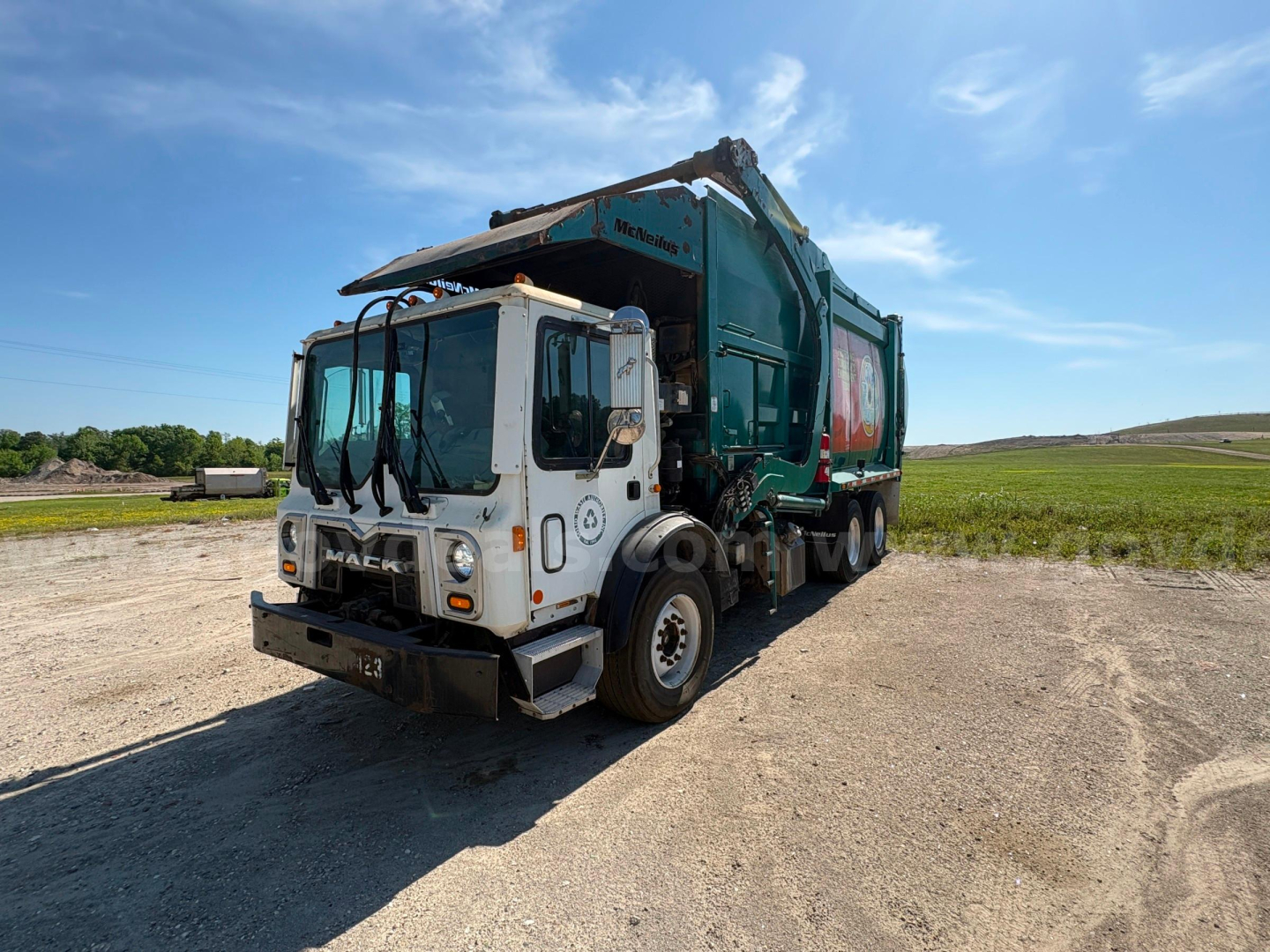 Garbage truck front-left view showing Mack MRU613 with dump body and graphic decals, white cab, green rear hopper, side mirror arrays, on dirt lot.