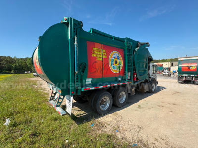 Rear and side view of a green Mack MRU613 refuse truck with text stickers and hazard emblem on the side, cargo body, wheels, and towing hitch visible.