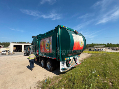 Rear three-quarter view of a green 2013 Mack MRU613 garbage truck with rear compactor, stickers, and dual rear axles.
