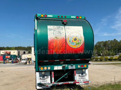 Rear view of a green garbage truck with reflective warning decals and a circular yellow-green logo; license plate area and bumper with white panels.