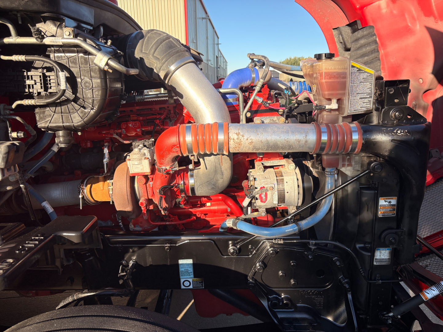 Kenworth T680 engine bay with red engine, exhaust pipe, hoses, and coolant reservoir visible.