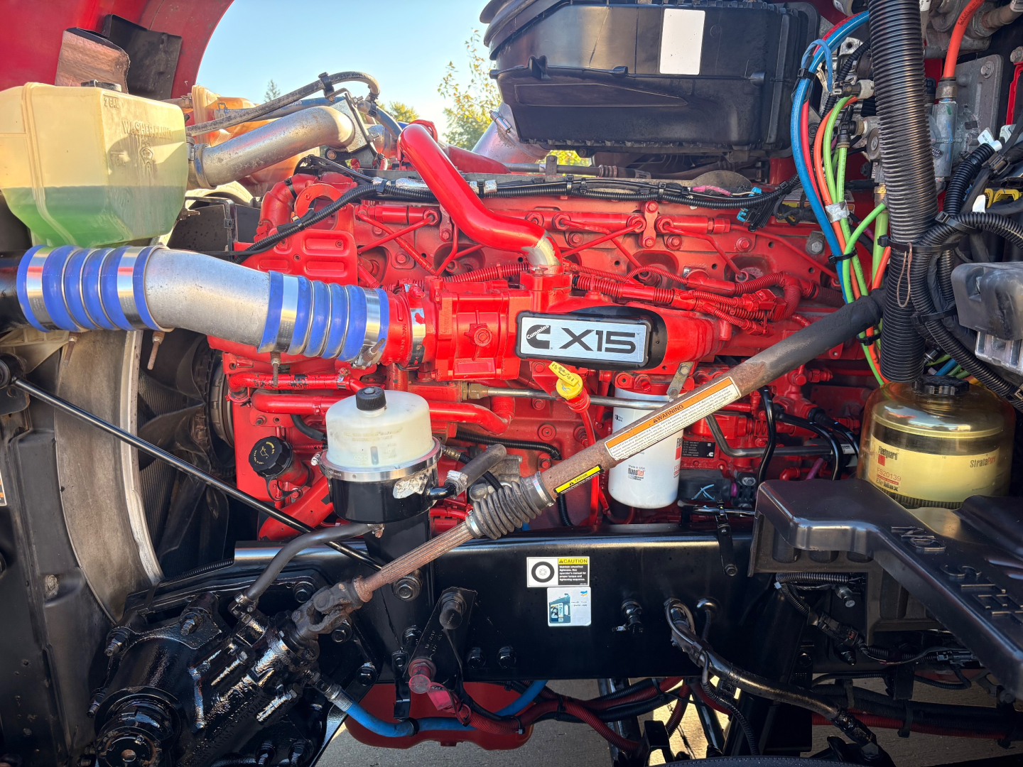 Kenworth T680 engine bay with red Cummins engine, EXIS badge, blue silicone hoses, and various fluid reservoirs visible.