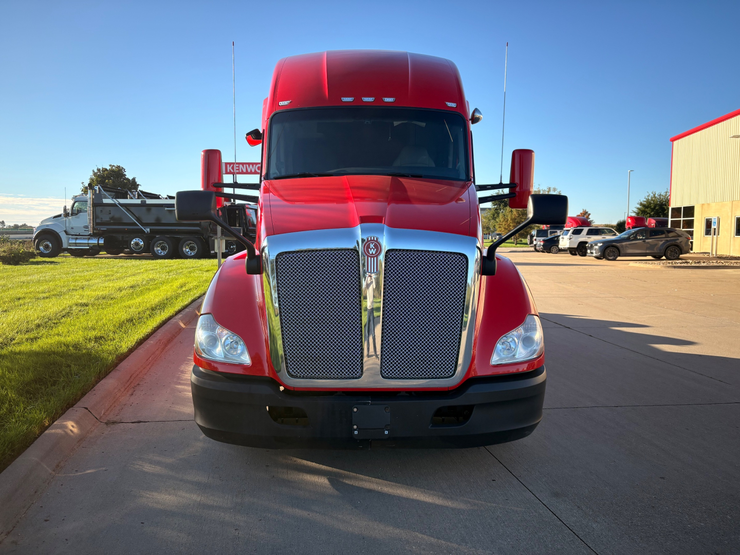 Truck front view of a red 2022 Kenworth T680 with chrome grille, large side mirrors, and KW emblem; clear sky, dealership lot in background.