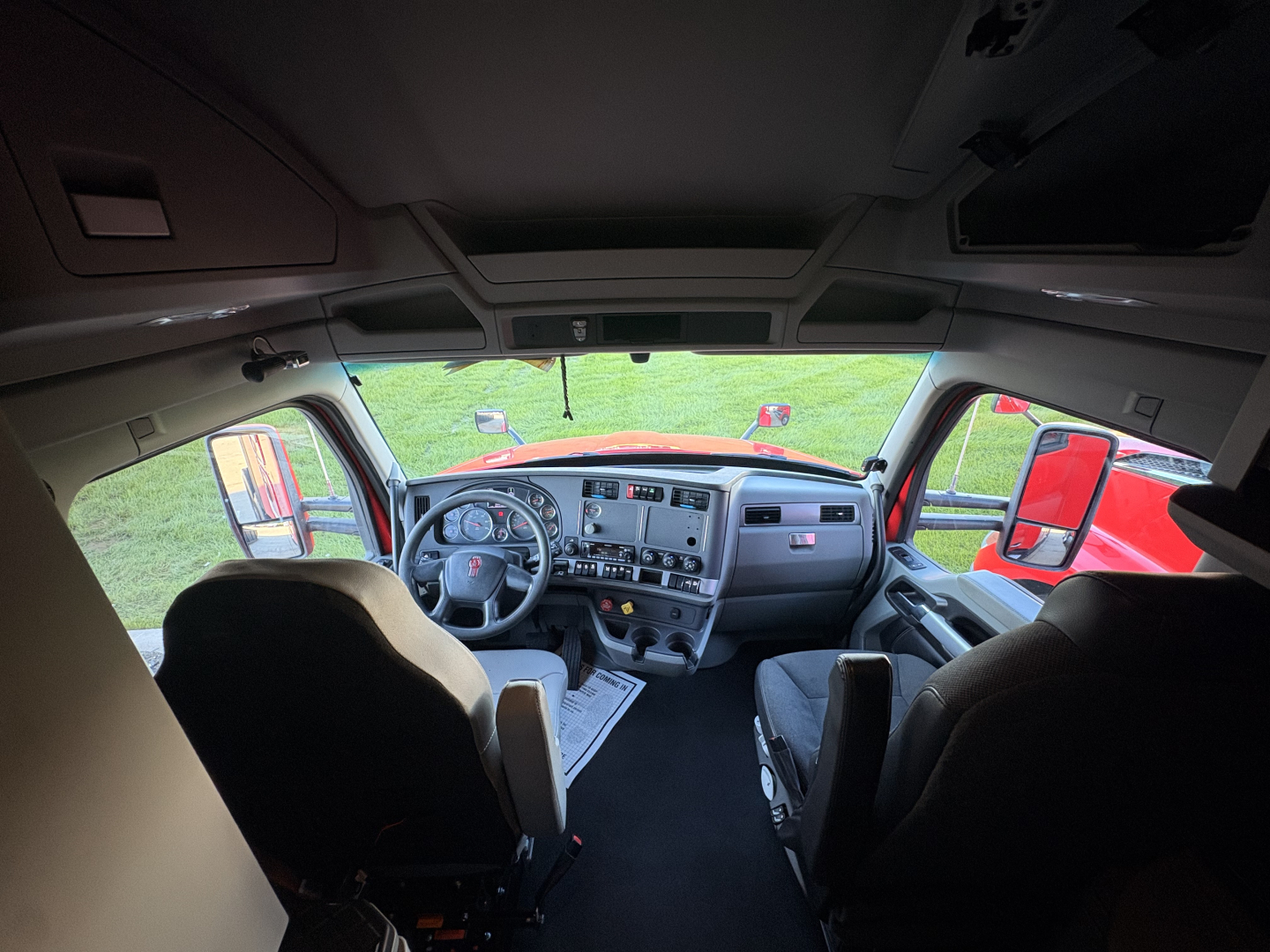 Interior of a Kenworth T680 cab, view from back seat showing steering wheel, dashboard, and driver/passenger seats.