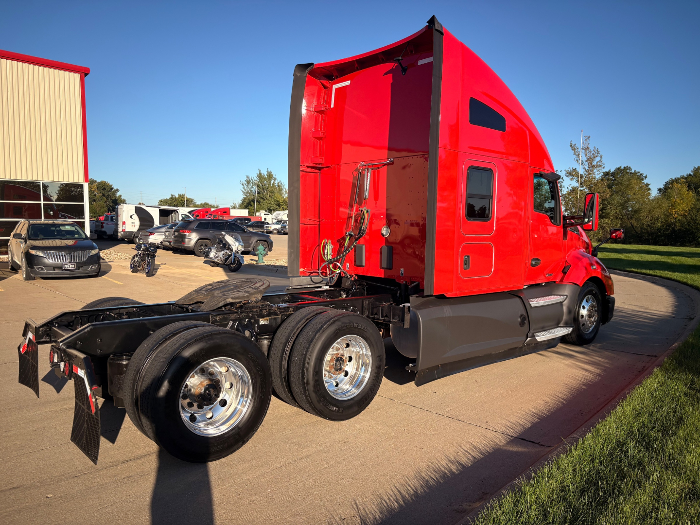Kenworth T680 sleeper cab truck in red, side view showing rear tractor frame and dual rear axles, with parked vehicles in background.