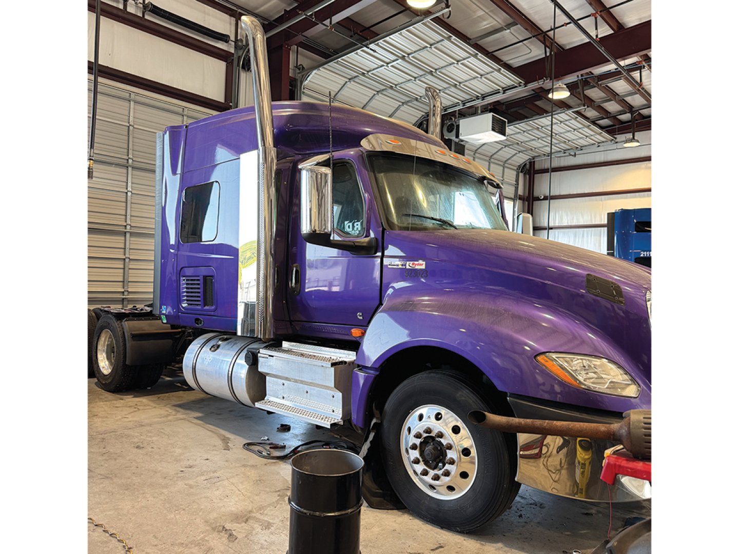 Truck cab in purple with chrome exhaust stacks showing interior of a sleeper tractor in a metal warehouse.
