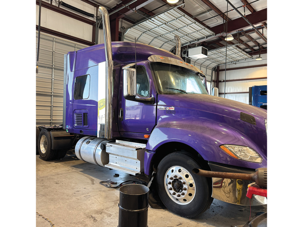 Truck cab in purple with chrome exhaust stacks showing interior of a sleeper tractor in a metal warehouse.