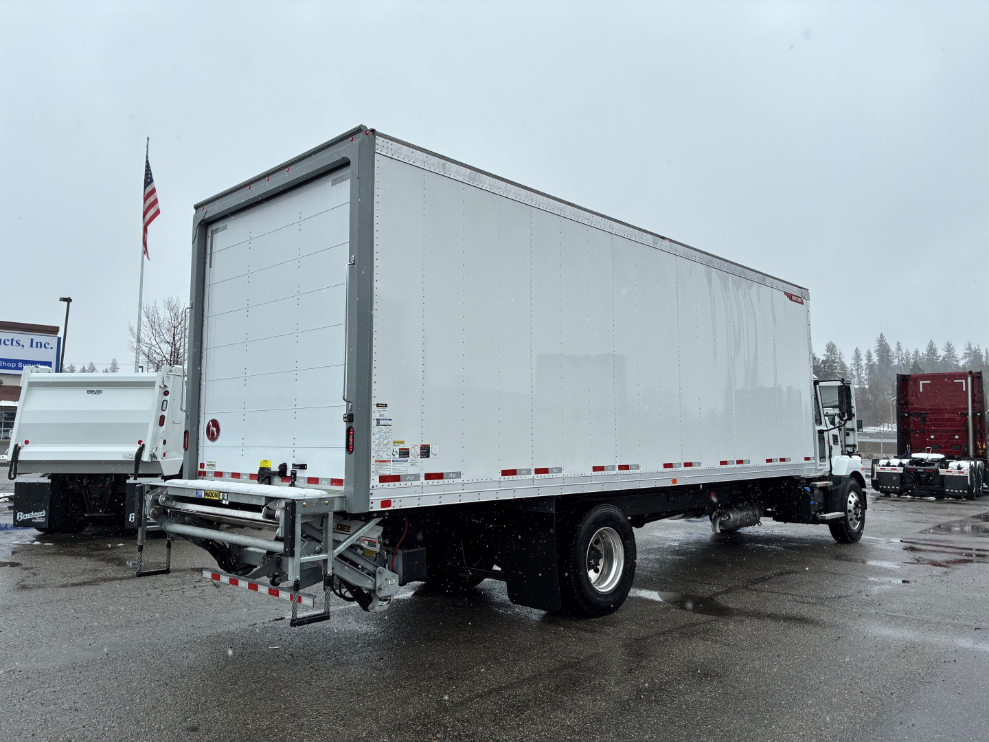 Box truck with a long rectangular cargo area and rear roll-up doors, Mack MD6 chassis visible in a dealership lot.